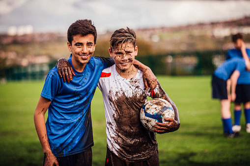 Teenage boys looking at the camera smiling after their soccer game. Both covered in mud. Other players can be seen blurred in the background.