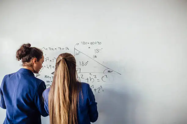 Two teenage school girls standing in front of  a large whiteboard side by side solving a mathematics equation on the board. Back view