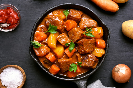 Beef meat stewed with potatoes and carrots in cast iron pan on dark background, top view