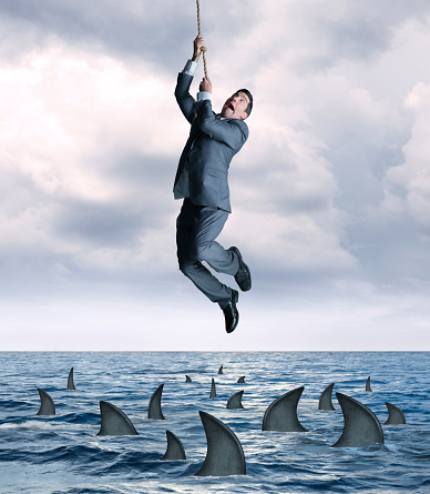 A businessman hangs on for dear life from a rope that dangles above a group of circling sharks in the ocean.  He looks up as the fear from falling into the dangerous waters grips his face.