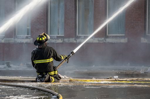 Saint John, New Brunswick, Canada - December 10, 2016. Fighting A Large Structure Fire. A fireman kneels in the middle of an icy road as he sprays water towards a brick building.