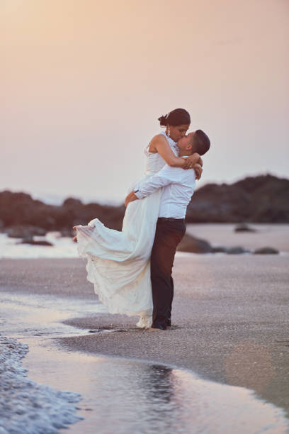 Happy kissing groom and bride Happy kissing groom and bride stand on ocean coastline in sunset light. Exotic wedding on beach island with pink sky background central america photos stock pictures, royalty-free photos & images