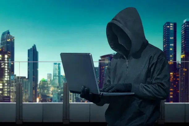 Hooded hacker with mask stealing information with laptop in the building terrace