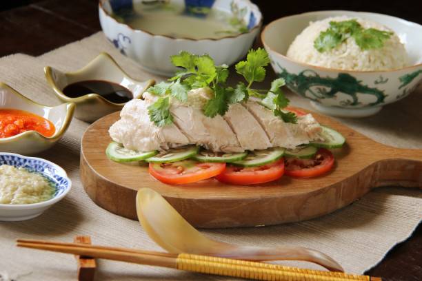 Singapore's Hainanese Chicken Rice Famous Singaporean dish of poached chicken with steamed rice cooked in the chicken stock; accompanied with sauces of garlic ginger, chili, and soy. Also served with the broth. The chicken meat is served on a wooden serving block while the rice and the broth are on vintage bowls. johor photos stock pictures, royalty-free photos & images