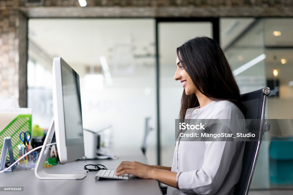 Smiling young businesswoman typing on computer Portrait of young Latin-American businesswoman wearing white shirt sitting at table in office, typing on computer and smiling Indian Ethnicity Stock Photo