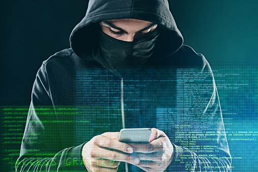 Shot of a computer hacker using a smartphone while standing against a dark background