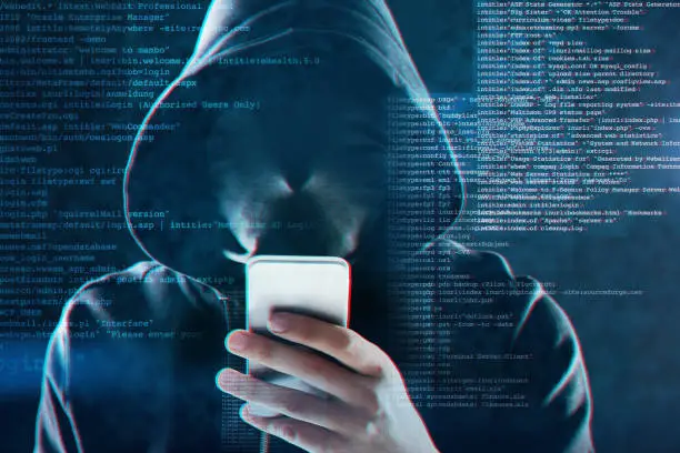 Shot of an unidentifiable computer hacker using a smartphone while standing against a dark background