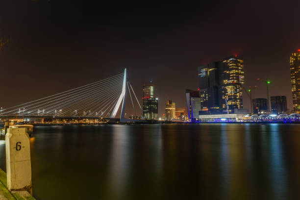 Rotterdam, the Netherlands, cityscape by night against a cloudy sky with the Erasmus bridge, a bollard close-up and illuminated buildings in the background. The bridge is a combined cable-stayed and bascule bridge in the centre of Rotterdam and it connects the northern and southern parts of this city. It takes its name from Erasmus of Rotterdam, a famous christian renaissance humanist. The project of the Erasmus bridge was made by the architect Ben van Berkel and it was built between the 1989 and 1990. The scene is calm and peaceful because the photo was taken around midnight, therefore there are no boats and no people.