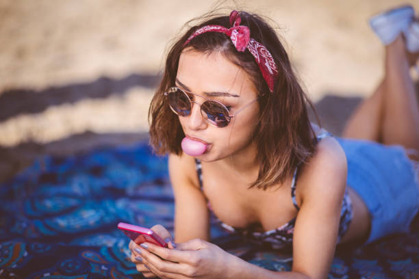 Young beautiful woman texting and blowing a bubble gum Young beautiful woman checking social media on her smart phone while blowing a bubble gum lying at the beach girls laying on beach stock pictures, royalty-free photos & images