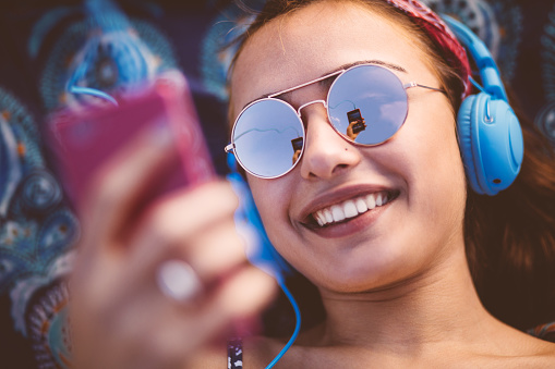 Beautiful young hipster woman wearing sunglasses and headphones, listening to music, texting and smiling