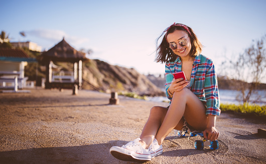 Attractive young woman sitting on skateboard and text-messaging next to the sea