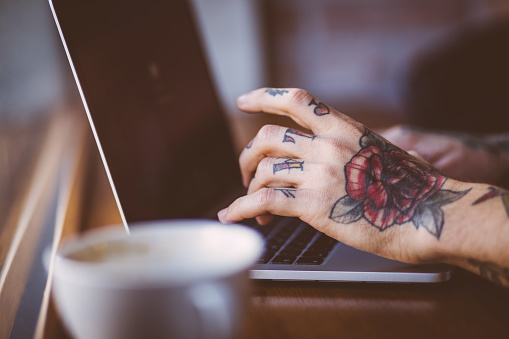 Close up of cool young guy's hands with tattoos typing on a laptop, working and chatting online with coffee in the background