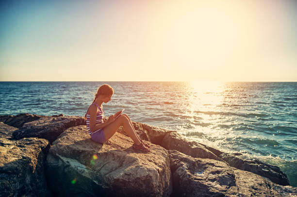 Little girl sitting alone on the rocks on tuscan beach. The girl is playing with tablet.\n