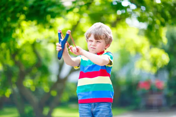 Funny little kid boy shooting wooden slingshot against green tree background. Child having fun in summer.