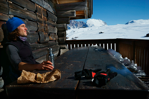 Single man wearing sports clothing and sitting in front of a wooden hut having a lunch break. Scenic winter landscape in the Dolomite Alps. Male winter sports athlete pausing in scenic alps landscape. Sunshine and clear sky on the Seiser Alm in Italy. Snow covered mountain range. XXXL (Sony Alpha 7R)