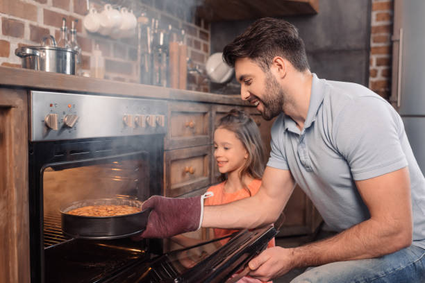 улыбаясь отец и дочь принимая торт из духовки - hot baking стоковые фото и изображения