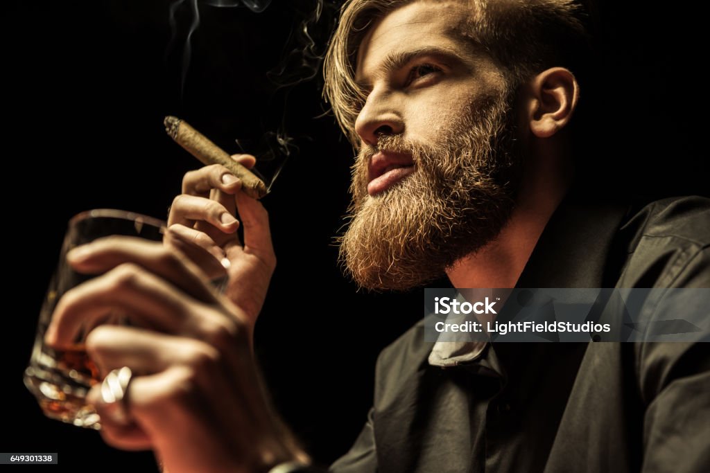 Handsome bearded man holding glass of whisky and smoking Handsome bearded man holding glass of whisky and smoking cigar on black Men Stock Photo