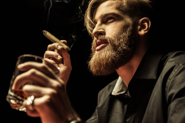 Handsome bearded man holding glass of whisky and smoking Handsome bearded man holding glass of whisky and smoking cigar on black man cigar stock pictures, royalty-free photos & images