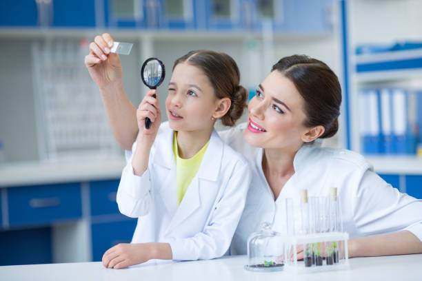 Mujer maestra y niña científicas estudiantes mirando diapositiva de microscopio de vidrio a través de la lupa - foto de stock