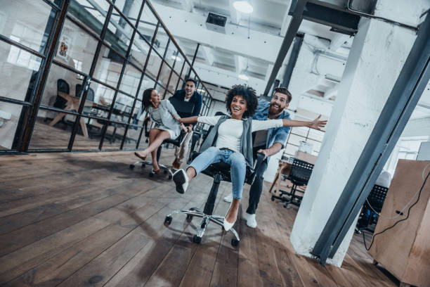 Work hard play hard! Four young cheerful business people in smart casual wear having fun while racing on office chairs and smiling office competition stock pictures, royalty-free photos & images