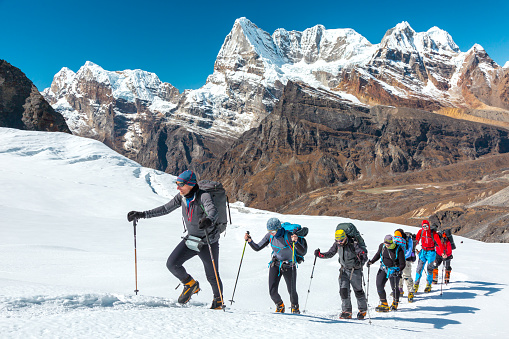 Adventurous People of variety of Age Origin and Ethnicity making Climb in high Altitude Mountains walking on Glacier with heavy Snow using trekking Poles and other Gear