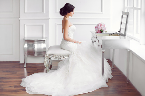 Wedding preparation. Beautiful young bride in white wedding dress indoors. Luxuty model looking at mirror sitting on chair with brides bouquet like at home in studio room with big window. Girl shows wedding fashion in decorated bright chic interior, high key. Side view.