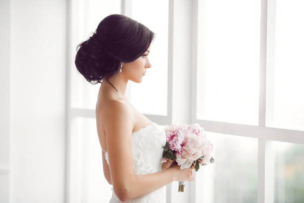 Stunning young bride holding bouquet Beautiful bride. Wedding hairstyle make-up luxury fashion dress and bouquet of flowers. Young attractive multi-racial Asian Caucasian model like a bride against white room with big window looking away. Side view bridal hair stock pictures, royalty-free photos & images