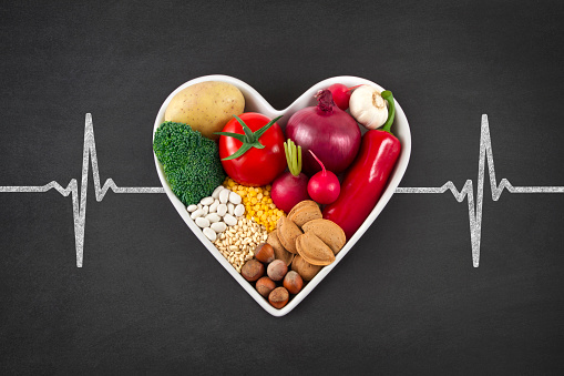 Heart health concept with related foods in white heart shaped bowl. potato red peppers, broccoli, radish, red onion, garlic, dry beans, almonds, nuts, and other legumes were arranged in heart shape plate on blackboard with drawing of pulse trace.