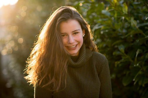 Outdoors portrait of a teenage girl with curly long brown hair in front of some leaves. She is looking happy at the camera wearing a green pullover.
