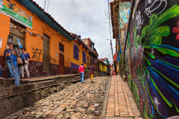 Bogotá, Colombia - Tourists, On The Narrow, Colorful, Cobblestoned Calle del Embudo In The Historic La Candelaria District Bogota, Colombia - July 20, 2016: To the left of the image, a tour guide explains some of the many legends of Colombia, on the narrow, cobblestoned Carrera Segunda or Second Avenue, in the historic district of La Candelaria. He is carrying an Arhuaca Mochila, a bag originally made by the Arhuaca tribe of the Sierra Nevada. Bogota the capital city of Colombia, was founded in 1538, in this area, by the Spanish Conquistador, Gonzalo Jiménez de Quesada. Not much seems to have changed in the last 470 years on this street, which was constructed when people travelled mainly on horseback. Many walls in the area are painted with either street art, or legends of the pre Colombian era, in the vibrant colours of Latin America. A small group of tourists listen to the guide. The sky is overcast; it will probably rain shortly. Photo shot in the morning sunlight, on a cloudy day. Horizontal format. Copy space. calle del embudo stock pictures, royalty-free photos & images