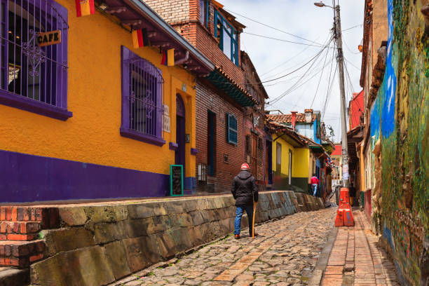 Bogotá, Colombia - Local Colombians Walk Through The Narrow, Colorful, Cobblestoned Calle del Embudo In The Historic La Candelaria District Bogota, Colombia - July 20, 2016: The narrow Carrera Segunda in the historical La Candelaria District of the South American capital city of Bogota, in Colombia is not too crowded in the image, because it is 20th July, National Day, and a holiday in the Country. It is in this area that the City was founded in 1538, by the Spanish Conquistador, Gonzalo Jiménez de Quesada. Not much seems to have changed in the last 470 odd years on this narrow street, which was constructed when people travelled mainly on horseback. Many walls in the area are painted with either street art, or legends of the pre Colombian era, in the vibrant colours of Latin America. Some local Colombians are seen in the image. The sky is overcast; it will probably rain shortly. Photo shot in the morning sunlight, on a cloudy day. Horizontal format. Copy space. calle del embudo stock pictures, royalty-free photos & images