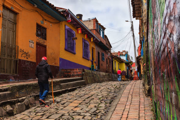 Bogotá, Colombia - Local Colombians Walk Through The Narrow, Colorful, Cobblestoned Calle del Embudo In The Historic La Candelaria District Bogota, Colombia - July 20, 2016: The narrow Carrera Segunda in the historical La Candelaria District of the South American capital city of Bogota, in Colombia is not too crowded in this image, because it is 20th July, National Day, and a holiday in the Country. It is in this area that the City was founded in 1538, by the Spanish Conquistador, Gonzalo Jiménez de Quesada. Not much seems to have changed in the last 470 odd years on this narrow street, which was constructed when people travelled mainly on horseback. Many walls in the area are painted with either street art, or legends of the pre Colombian era, in the vibrant colours of Latin America. Some local Colombian tourists are seen in the image. The sky is overcast; it will probably rain shortly. Photo shot in the morning sunlight, on a cloudy day. Horizontal format. Copy space. calle del embudo stock pictures, royalty-free photos & images