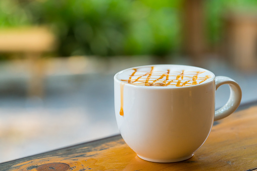 Hot Macchiato coffee with caramel in white cup on wood table by window light, copy-space background