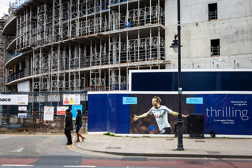 London, UK - February 22, 2017: two women, one of African and one of Asian ethnicity, walk along Tottenham High Road in London. Just behind them, the construction frame for Tottenham Hotspur FC's new 61,000 capacity stadium, can be seen. On the advertising hoarding parallel to the women, one Tottenham Hotspur's main players, Harry Kane, holds his arms out in celebration. Horizontal colour image with copy space.