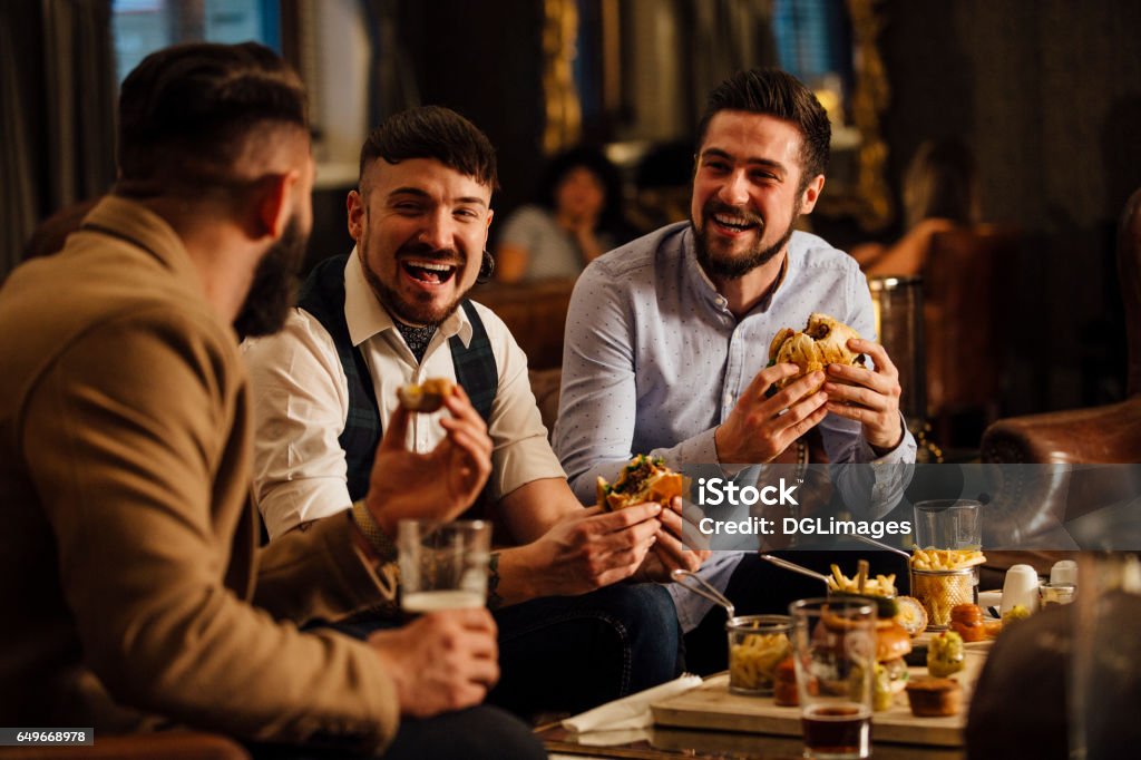 Pub Food And Drinks Three men are sitting together in a bar/restaurant lounge. They are laughing and talkig while enjoying burgers and beer. Friendship Stock Photo