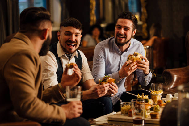 Pub Food And Drinks Three men are sitting together in a bar/restaurant lounge. They are laughing and talkig while enjoying burgers and beer. after work stock pictures, royalty-free photos & images