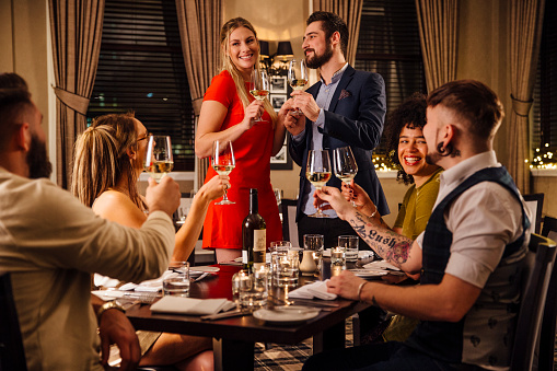 Friends are at a formal meal together. A couple are standing together at one end of the table, making a toast to which everyone is raising their glass.
