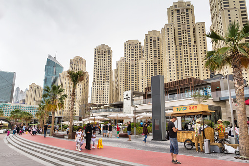 View of the new promenade on Dubai Marina with many restaurants and people that walking on pedestrian walkway. On background some hotels and apartments.