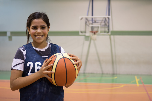 Young female basketball player holding ball and sitting on bench at indoors court and looking at camera portrait