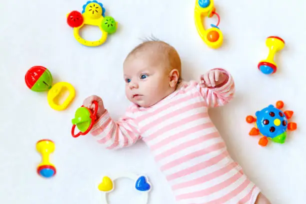 Cute adorable newborn baby playing with lots of colorful rattle toys on white background. New born child, little girl looking surprised at the camera. Family, new life, childhood, beginning concept