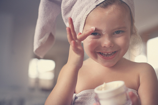 Cute little girl putting cream on her face. Looking at the camera.