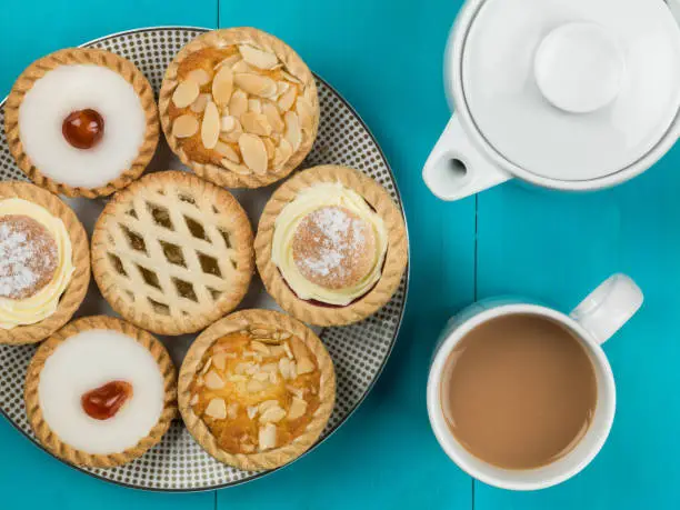 Plate of Assorted Individual Cakes or Tarts With a Pot of Tea Against a Blue Background
