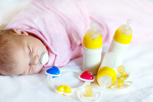 Cute newborn baby girl with nursing bottles, rattle and pacifier. Formula drink for babies. New born child, little girl laying in bed. Family, new life, childhood, beginning, bottle-feeding concept.