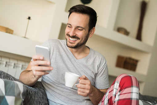 Young cheerful man is using mobile phone and reading text messages while enjoying his cup of coffee at home.