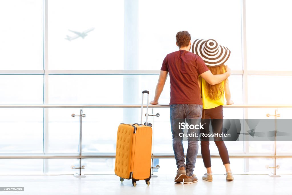 Couple at the airport Young couple in colorful t-shirts waiting with baggage near the window at the departure area of the airport during their summer vacation Couple - Relationship Stock Photo