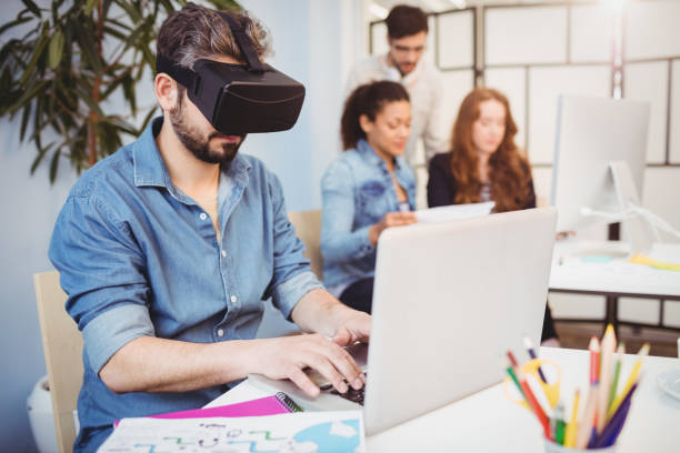 istock Businessman wearing virtual reality headset while using laptop against colleagues 649804134