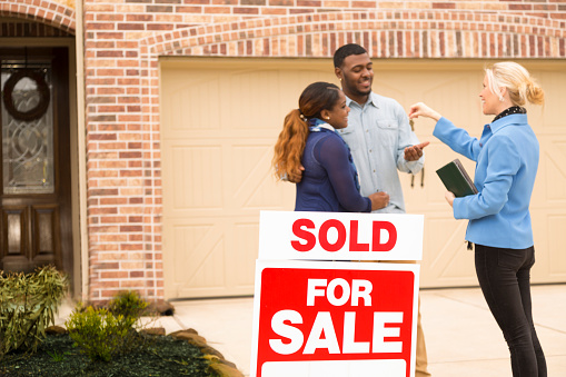 Excited African descent couple receives the keys to their first home real estate purchase from Real Estate Agent.  The happy young adults stand in front of brick home.