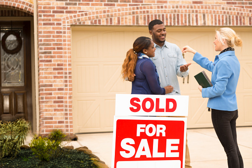 A family of four are standing outside of their new home. The sign in front of the house says, \