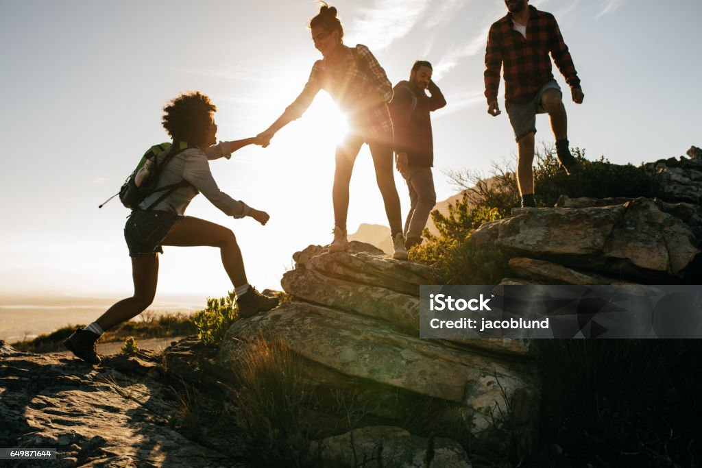 Young people on mountain hike at sunset Group of hikers on a mountain. Woman helping her friend to climb a rock. Young people on mountain hike at sunset. Hiking Stock Photo