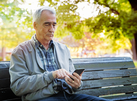 Senior man using mobile phone alone in a public park on a beautiful Autumn afternoon. He is frowning as he touches screen with one finger.
